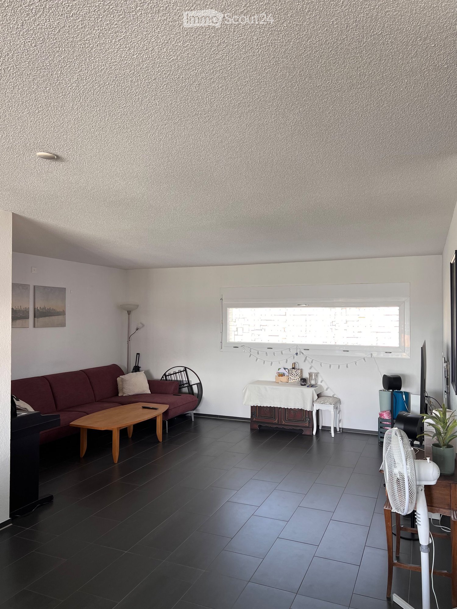 Living room with dark tiled floor, red sofa, wooden coffee table, small table with white cloth, fan, potted plant, and picture frames on the wall.