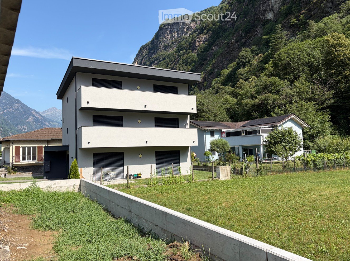 Modern two-story house with white exterior, large windows, and a balcony, situated in a green area with mountains in the background.