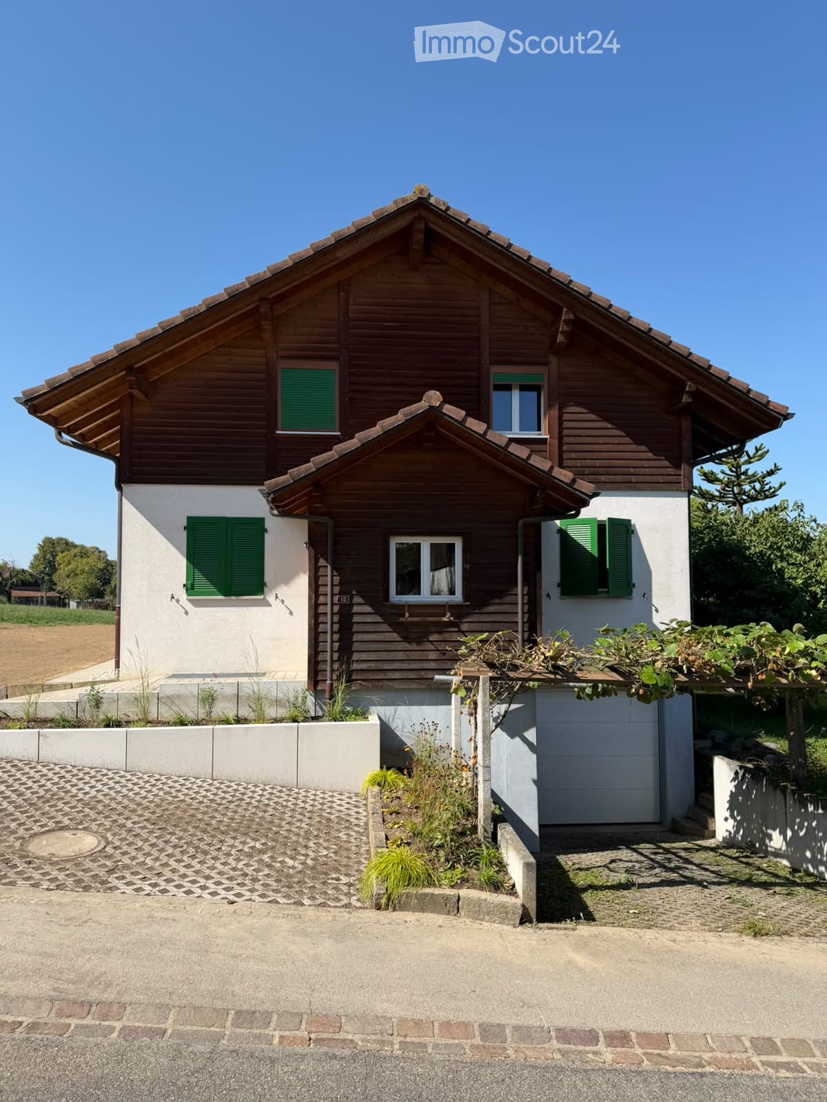 Two-story house with wooden siding, green shutters, and a raised ground floor. Features a garage and small garden area.
