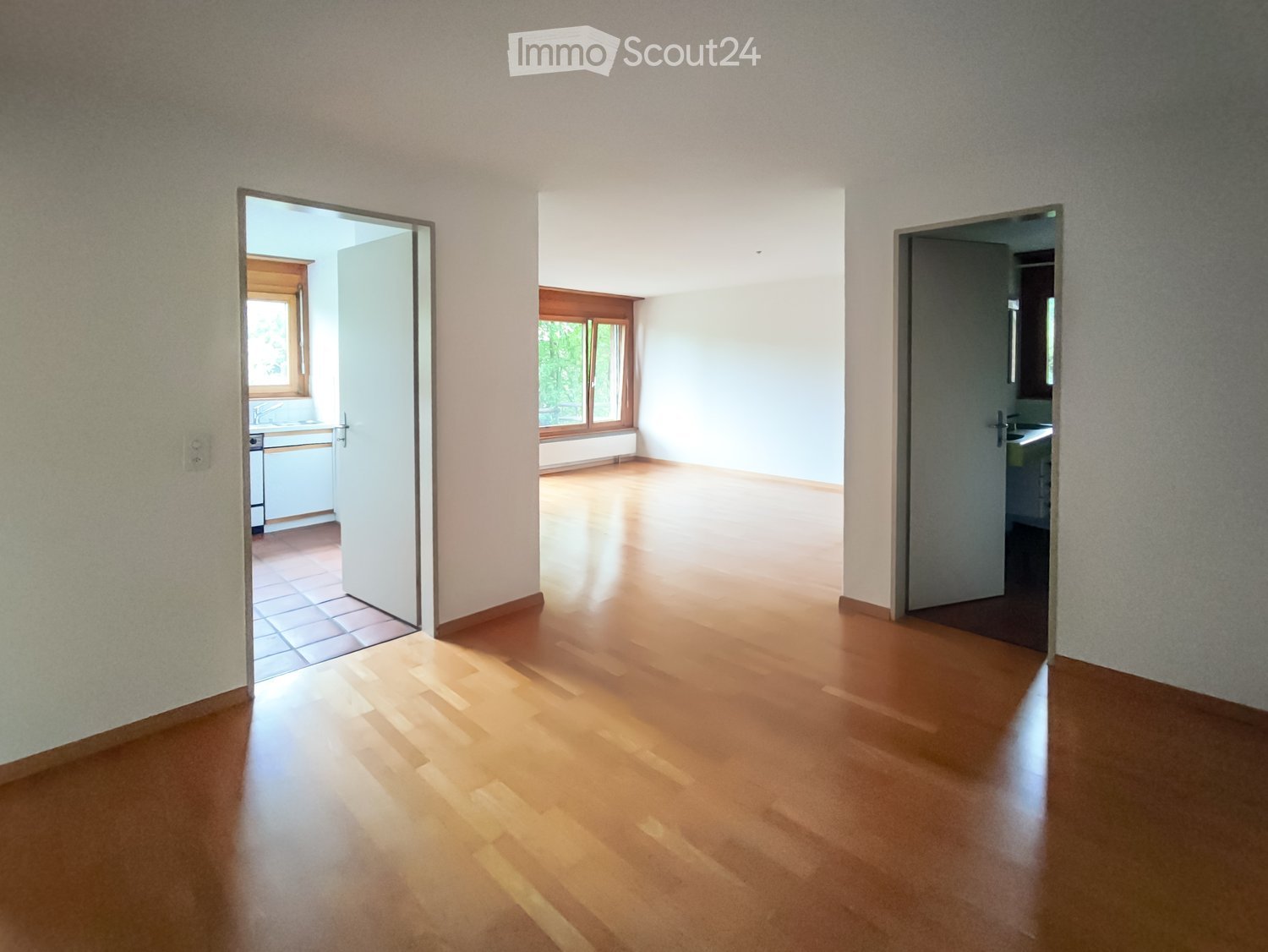Empty room with wooden flooring and white walls, featuring open doors leading to a kitchen and bathroom.