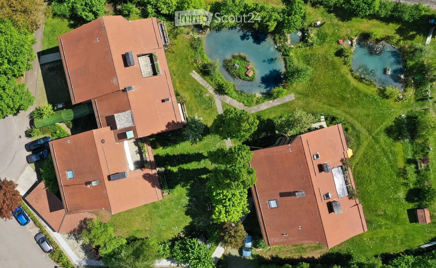 Aerial view of a residential house with a red-tiled roof, surrounded by a well-maintained garden, a pond, and several parked cars.