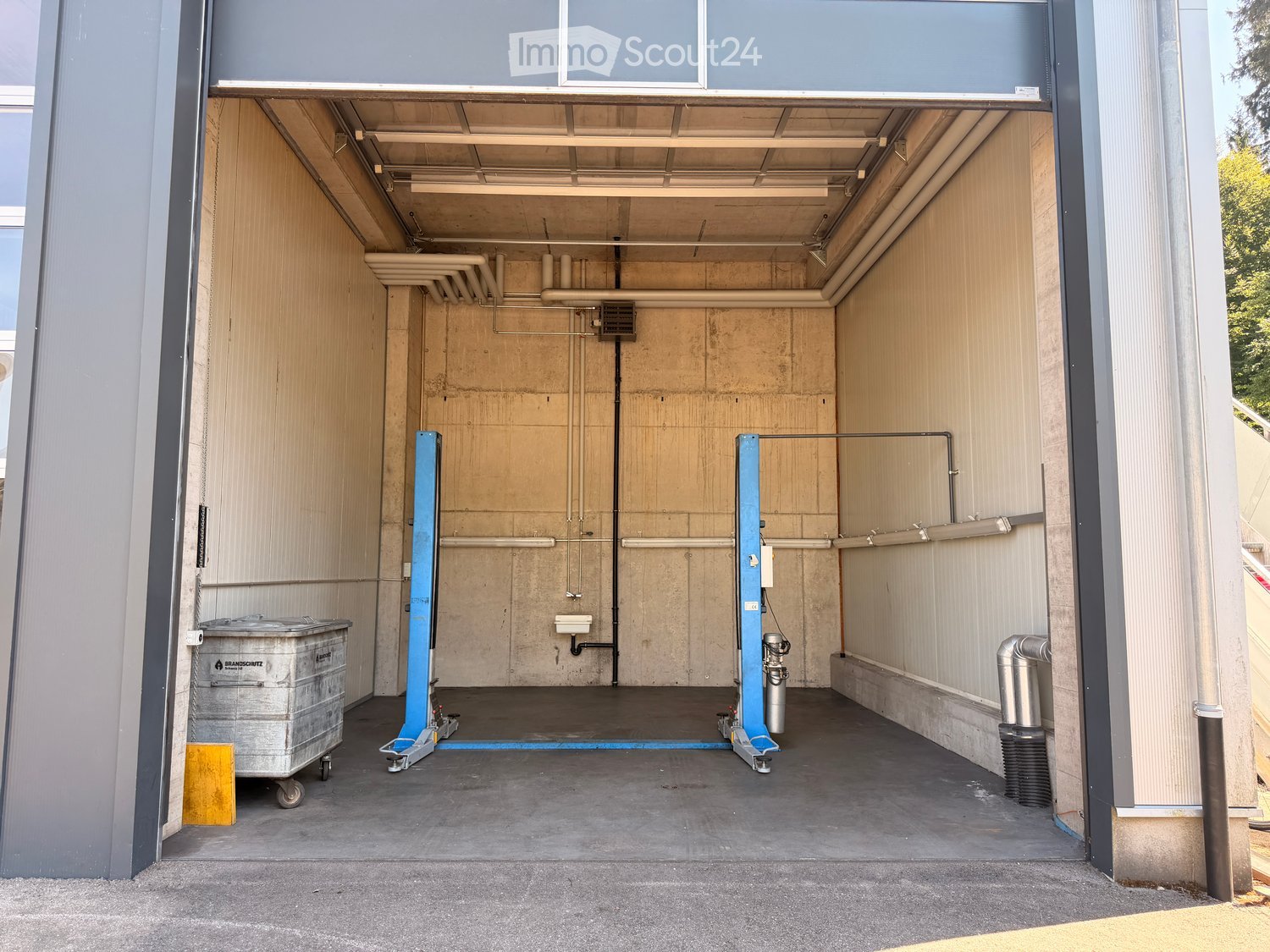 Open garage with concrete floor, metal walls, and industrial equipment including a blue lift and a trash bin.