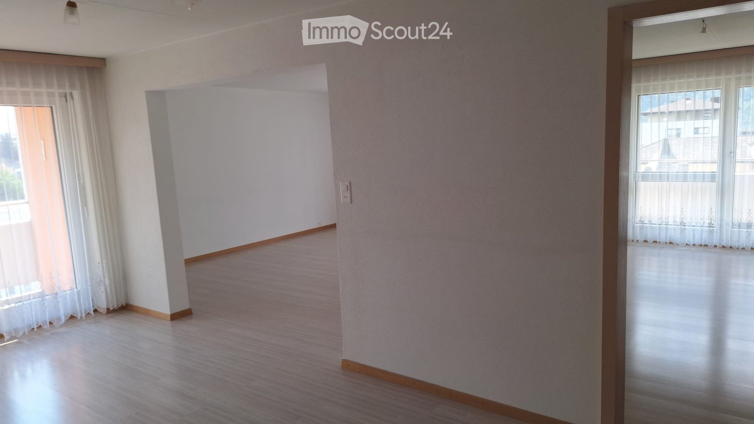 Empty room with light wooden flooring, white walls, and large windows with sheer curtains. No furniture or equipment present.