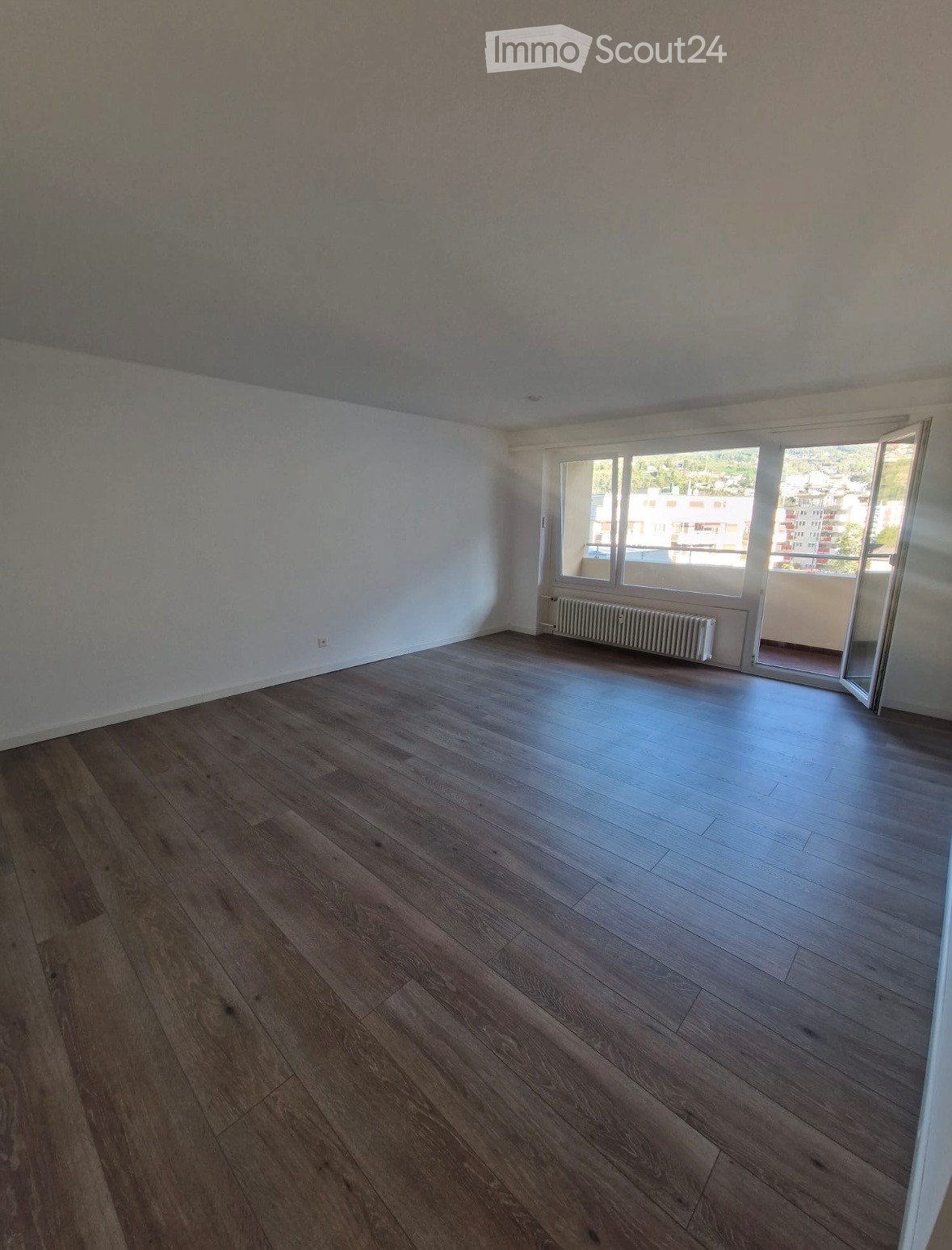 Empty room with wooden flooring, white walls, large window with view, radiator, and open sliding door leading to balcony.