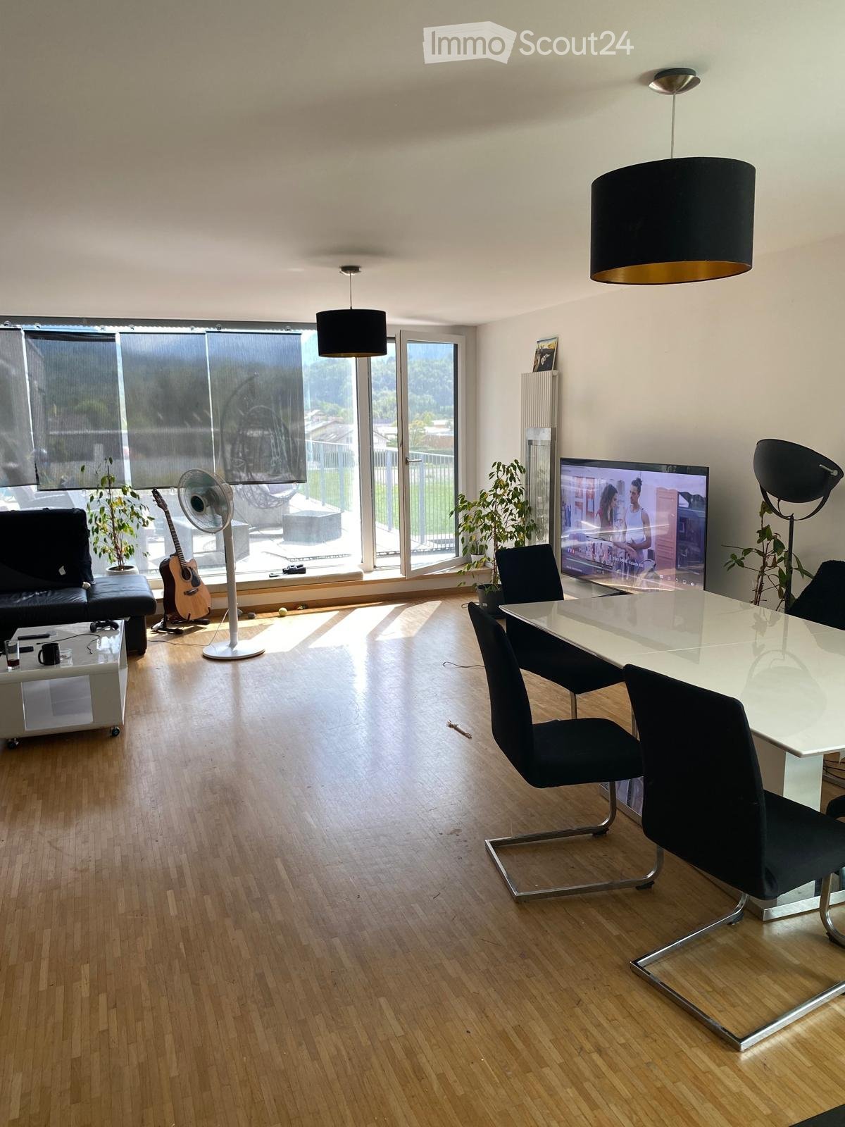 Modern living room with wooden flooring, large windows, a sofa, a coffee table, a TV, a guitar, a fan, and a dining area with a table and chairs.