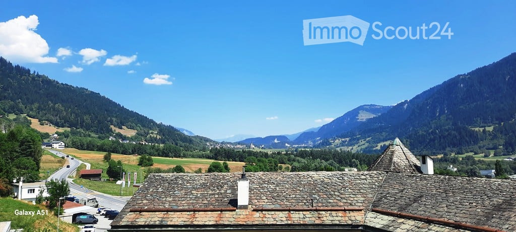 A picturesque view of a traditional stone-roofed building nestled in a lush, green valley surrounded by towering mountains under a clear blue sky.