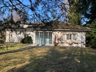 A single-story house with a white exterior, large windows, and a covered entrance. The roof is dark, and the surrounding area includes a lawn and some trees.