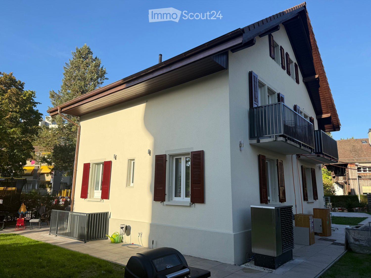 Two-story house with white walls, red shutters, and a balcony on the upper floor. The house has a well-maintained garden with a barbecue, a small fence, and outdoor furniture.