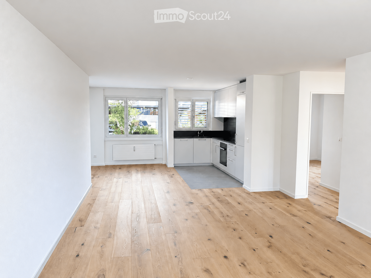 Bright, minimalist kitchen with white cabinets, a sink, and an oven, featuring light wooden flooring and large windows offering a view of greenery.