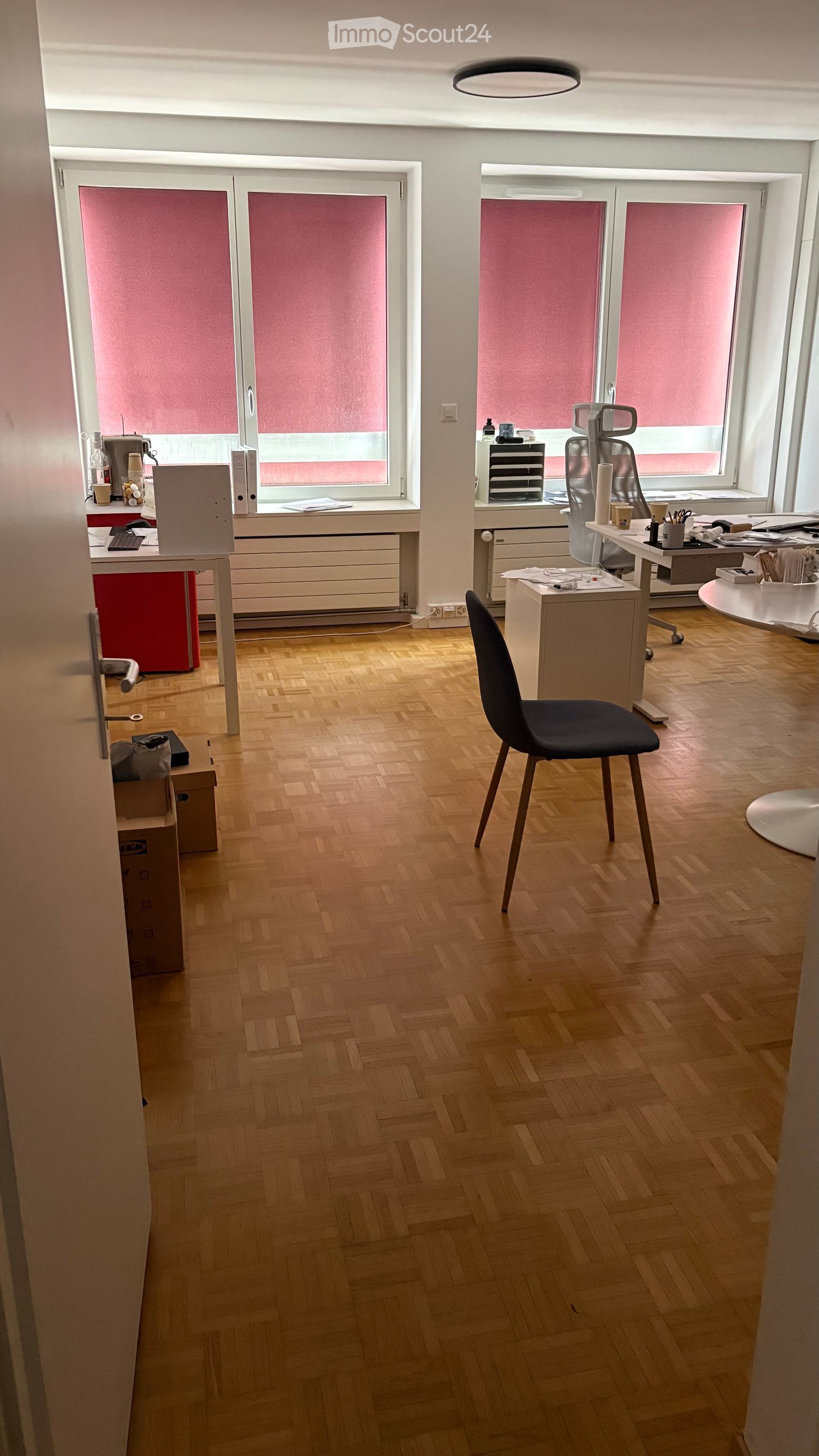 Office space with wooden herringbone flooring, large windows with red blinds, desks, chairs, and office equipment.