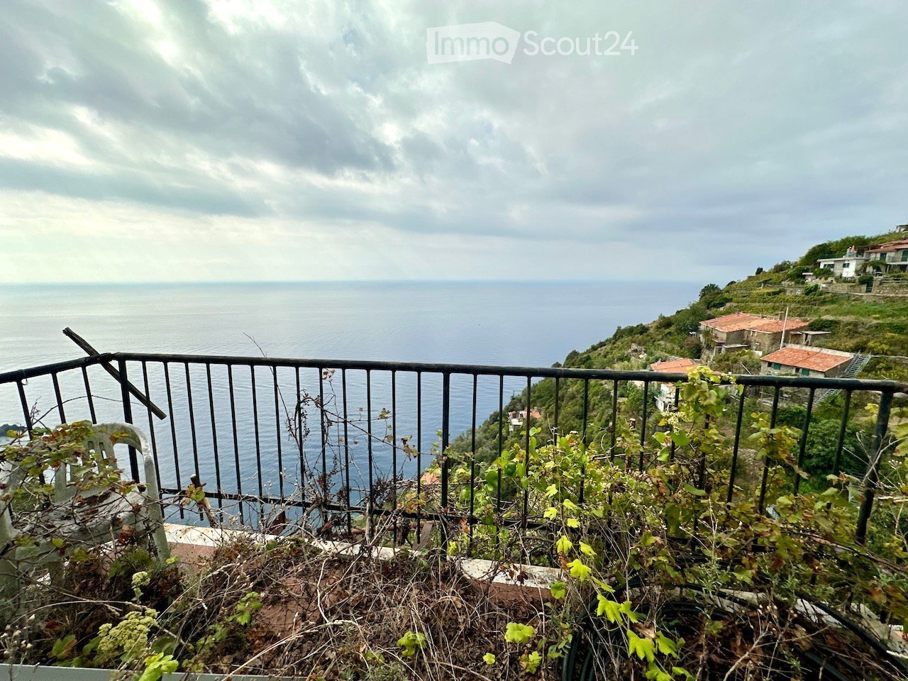 Balcony overlooking the sea with a metal railing, some overgrown plants, and distant houses on a hillside.