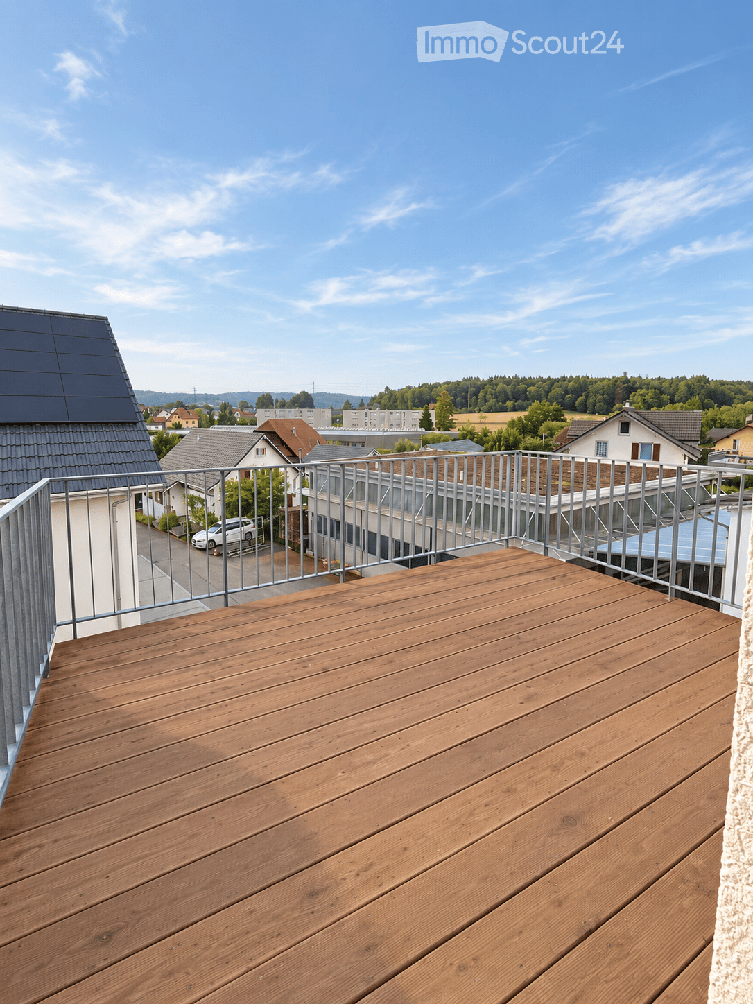 Wooden deck with metal railing, overlooking a suburban neighborhood with houses, trees, and a clear blue sky.