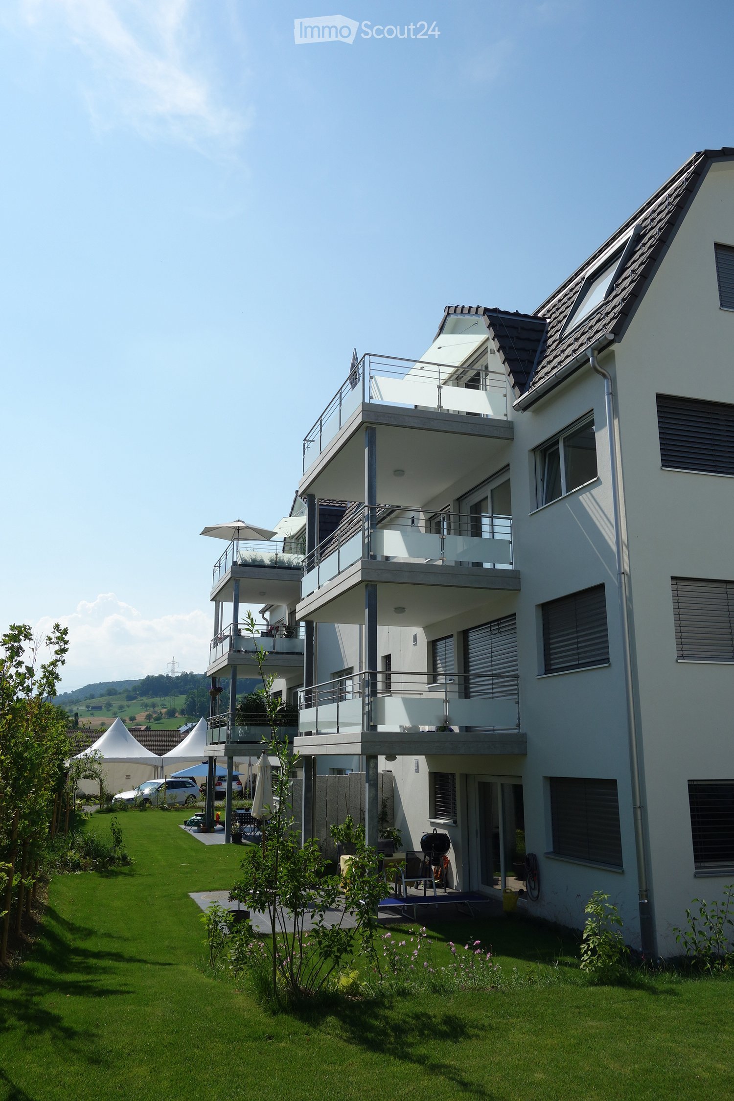 Modern two-story house with multiple balconies, large windows, and a well-maintained lawn with trees and shrubs.