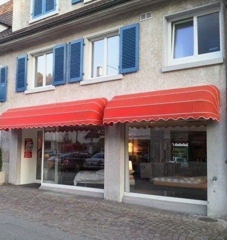A two-story building with blue shutters and red awnings over large windows. The ground floor has glass storefronts displaying interior furnishings.