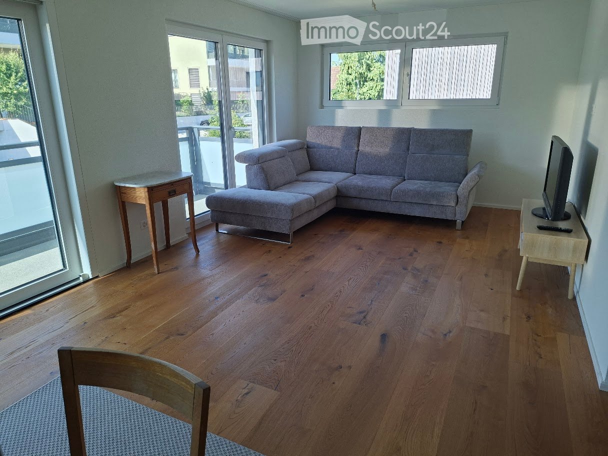 Modern living room with wooden flooring, large windows, and a grey sectional sofa. Includes a small wooden table, a television, and a wooden chair.