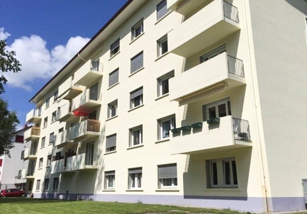 Multi-story residential building with multiple balconies and windows, light-colored facade, and a small lawn area in front.