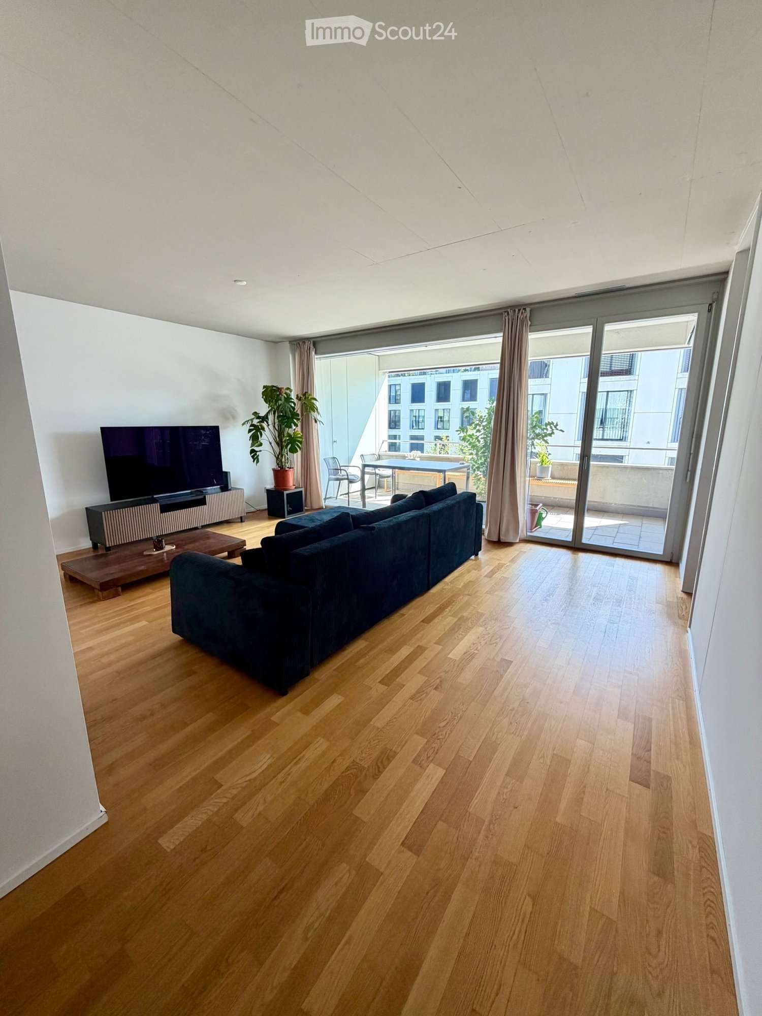 Modern living room with wooden flooring, large sofa, coffee table, TV, and sliding glass doors leading to a balcony with outdoor furniture.