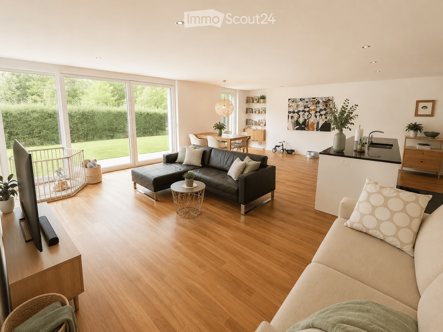 Open-plan living and dining area with wooden flooring, large windows, a black leather sofa, a beige sofa, a coffee table, a dining table with chairs, a kitchen counter, and various decorative items.