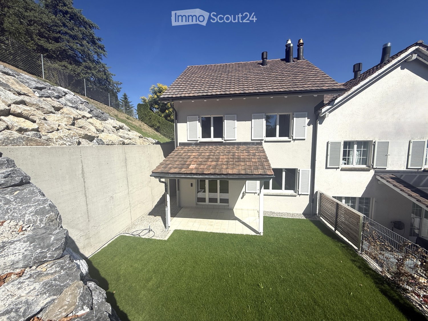 Two-story house with a tiled roof, multiple windows with shutters, and a small attached garage. The property includes a well-maintained lawn and a stone retaining wall.