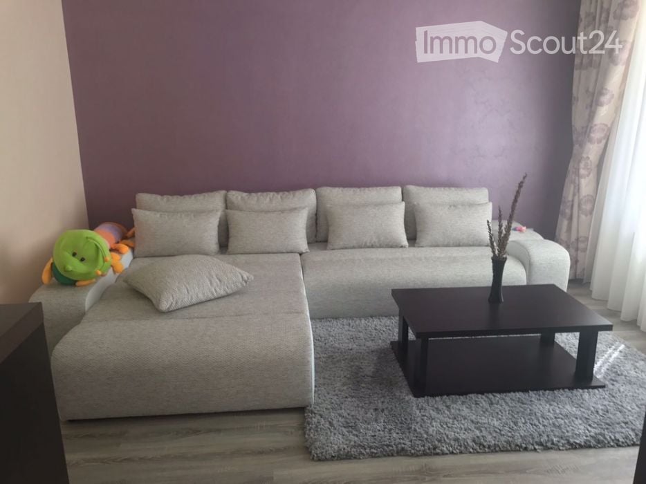 Modern living room with a grey sectional sofa, multiple pillows, a black coffee table with a vase, and a plush toy on the sofa. The room features wooden flooring and large windows with curtains.