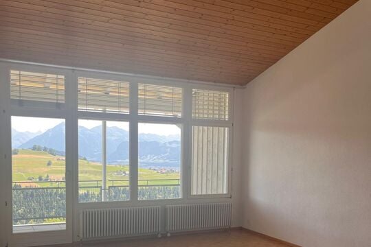 Empty room with large windows offering a scenic mountain view, wooden ceiling, and radiators.