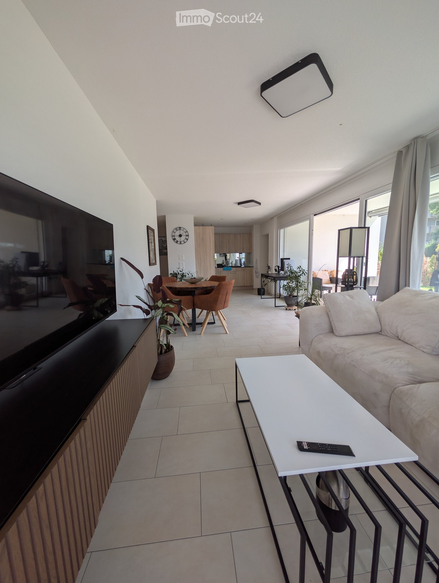 Modern living room with a beige sofa, white coffee table, TV stand, and a dining area with chairs and a table. The space features large windows and a sleek design.