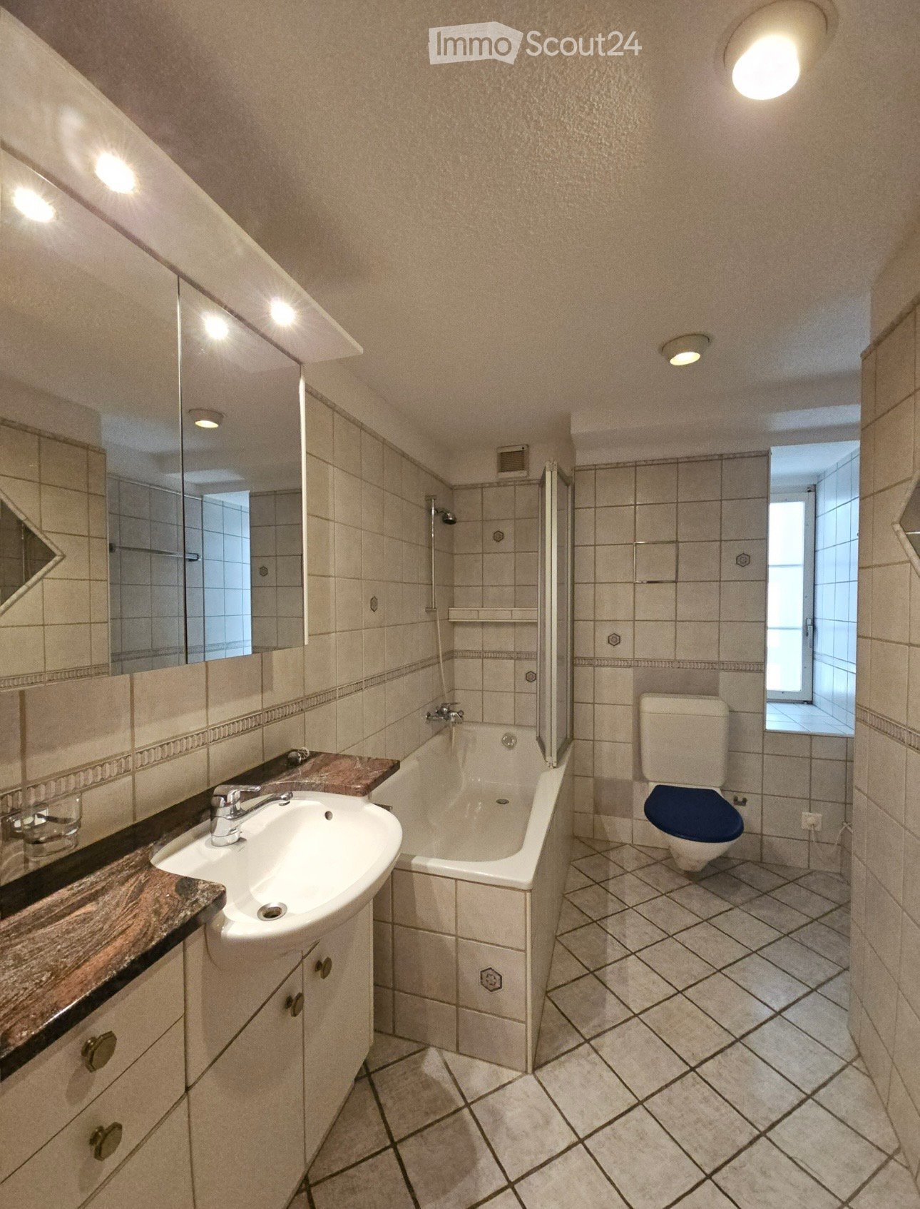 Bathroom with a white sink, bathtub, toilet, and mirror. The walls are tiled, and the floor is tiled with a diamond pattern. There is a window providing natural light.