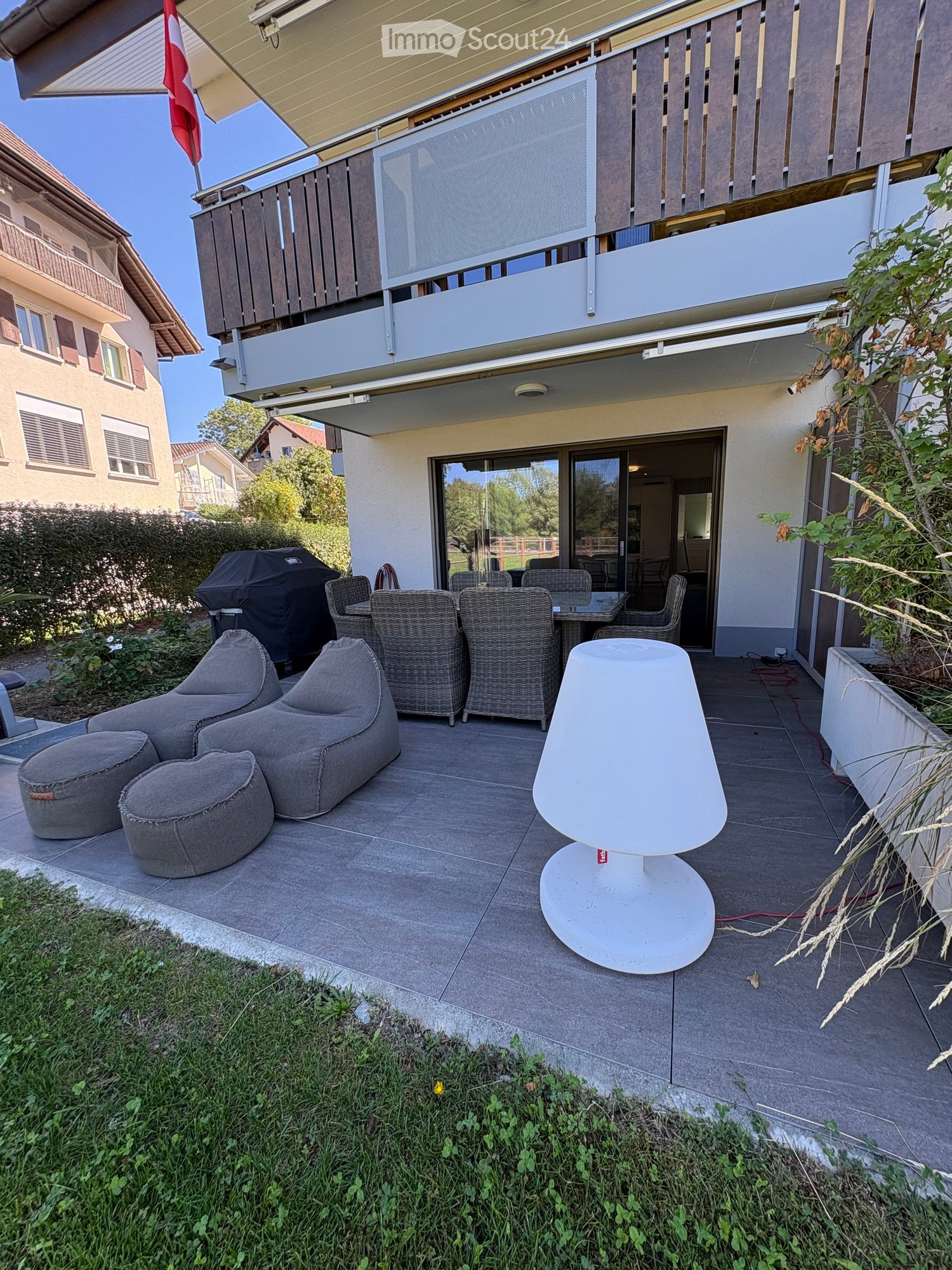 Outdoor patio with grey sofas, wicker chairs, a table, a white lamp, and a covered grill. The patio is tiled and has a view of the surrounding neighborhood.