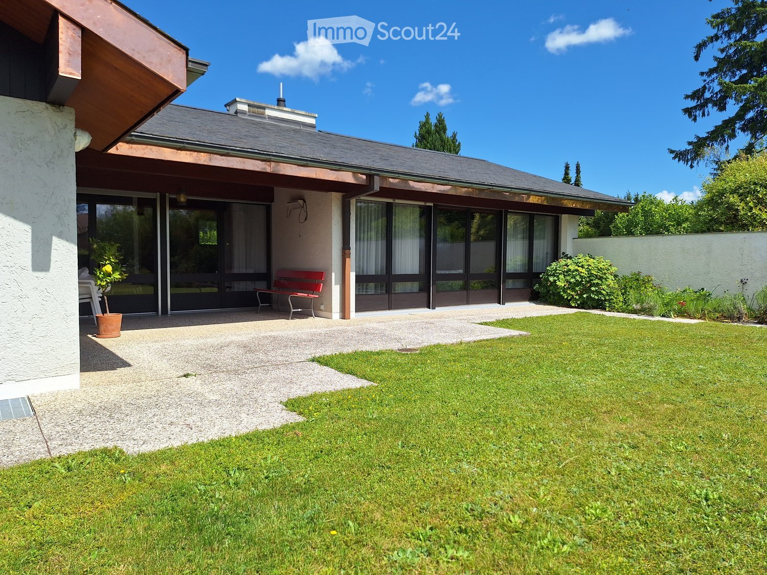 Well-maintained garden with a lawn, a small patio area, a red bench, and a potted plant near the house entrance.