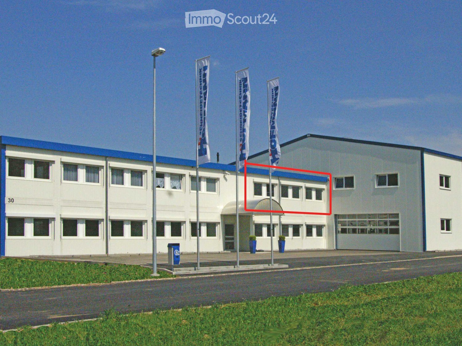 Two-story building with white walls, blue roof trim, multiple windows, and a large garage section. Flags are displayed on poles in front of the building.