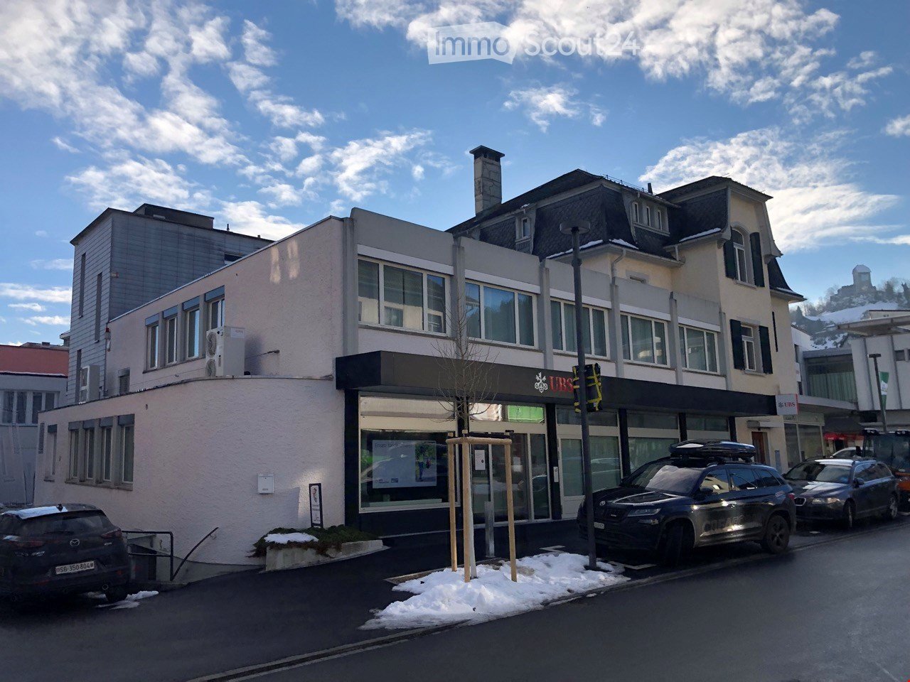 A multi-story commercial building with a storefront labeled "URS" and several parked cars in front, under a partly cloudy sky with snow on the ground.