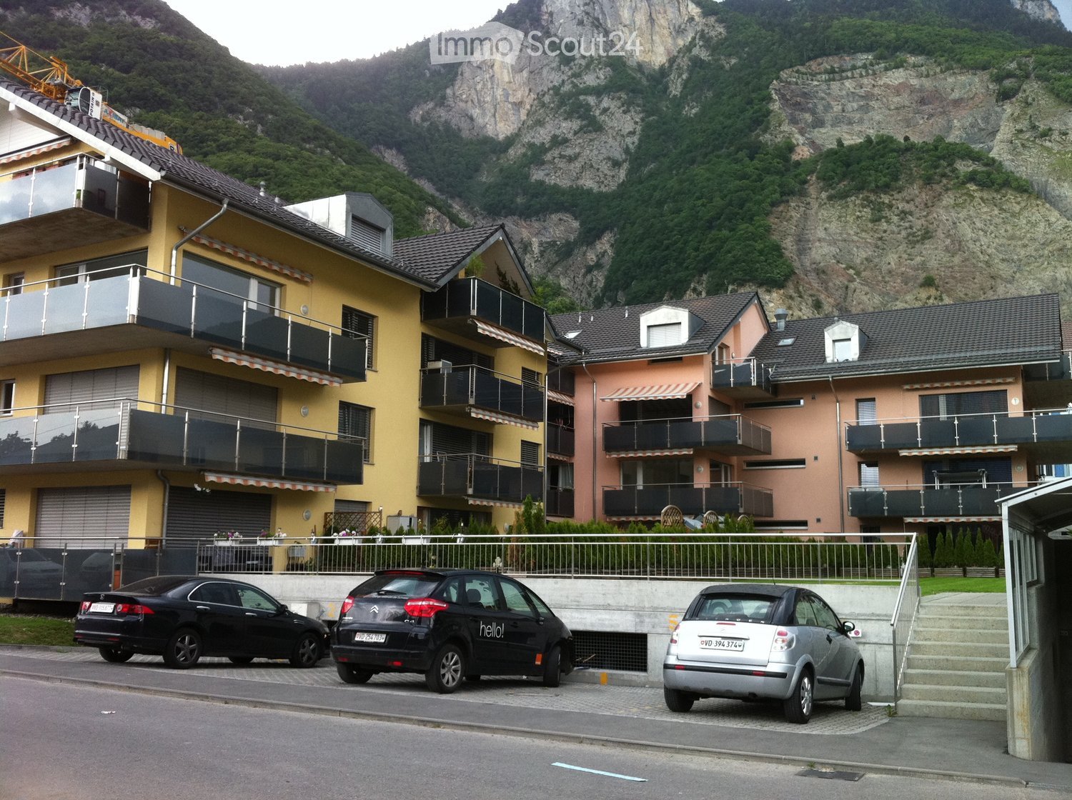 Multi-story residential building with balconies, surrounded by mountains, with parked cars in front.