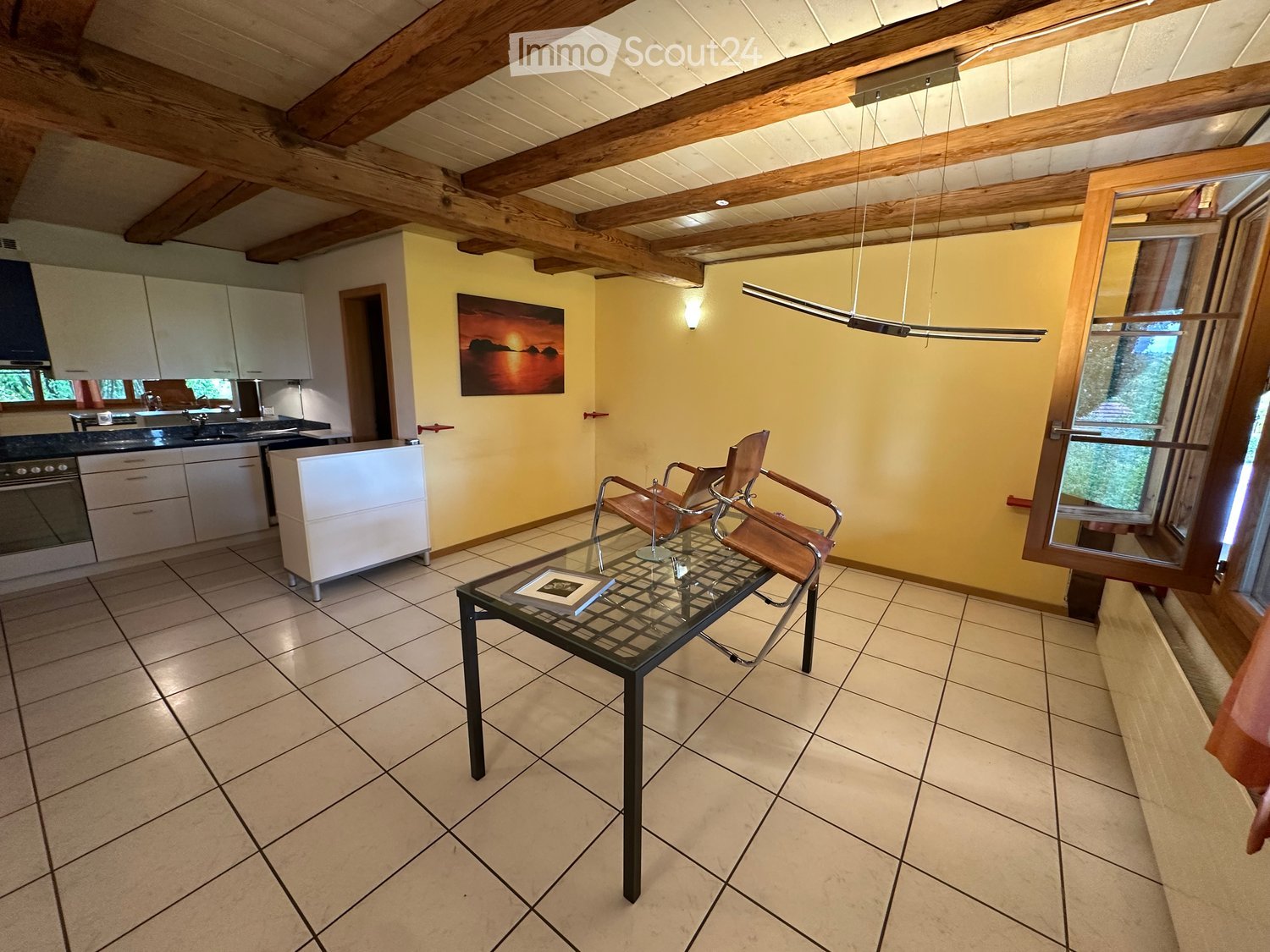 Open-plan living and kitchen area with exposed wooden beams, tiled floor, glass coffee table, two chairs, kitchen cabinets, oven, sink, and a painting on the wall.