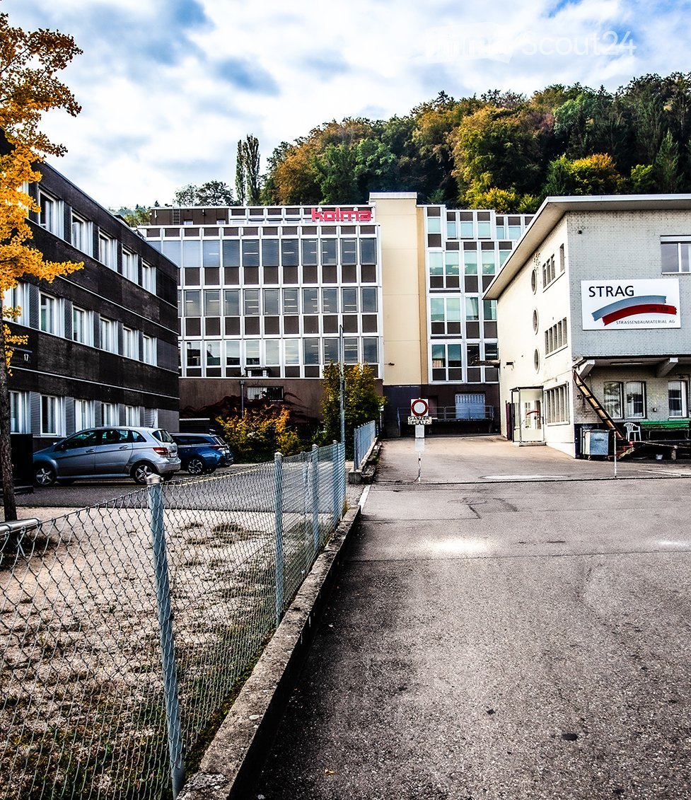 Multi-story industrial or commercial building complex with large windows, signage, and a fenced parking area.