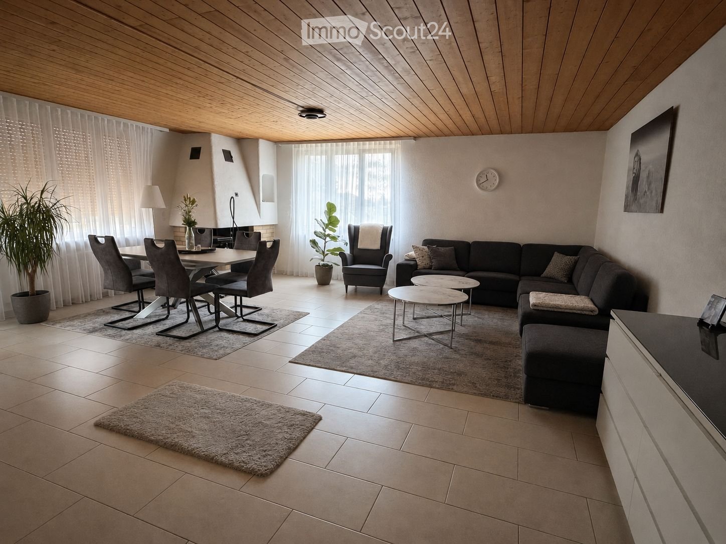Modern living room with a sectional sofa, coffee tables, dining area with chairs and table, large windows with curtains, and decorative plants.
