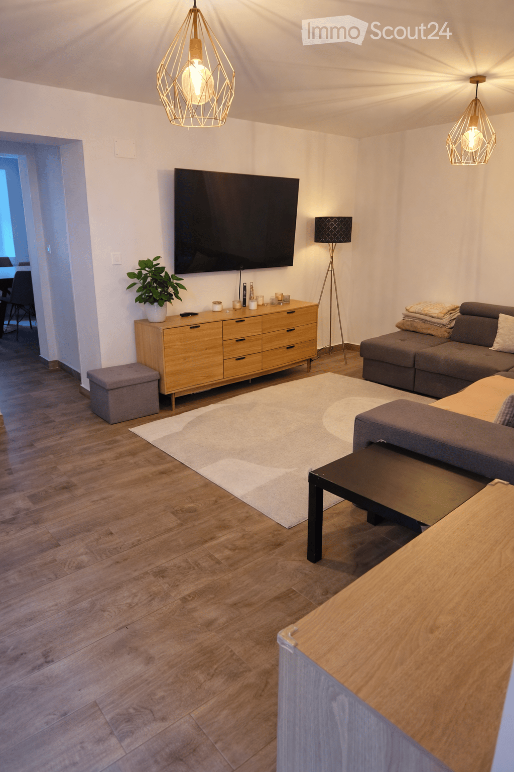 Modern living room with wooden flooring, a grey sofa, a coffee table, a television mounted on the wall, a wooden sideboard, a small stool, and decorative lighting.