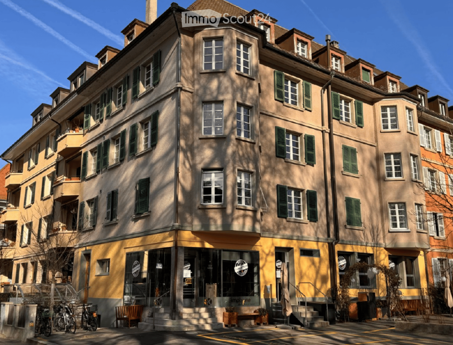 Multi-story residential building with beige and yellow facade, green shutters, and multiple windows. Features a storefront with glass doors and a small staircase leading to the entrance.