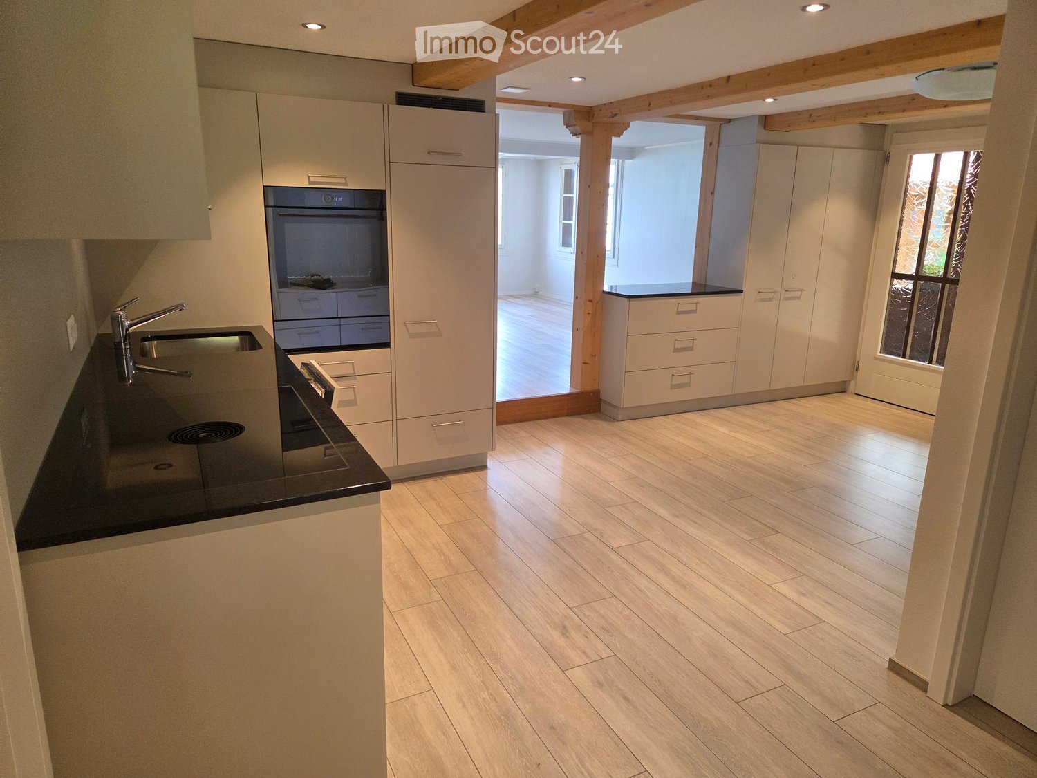 Modern kitchen with light-colored cabinets, black countertop, sink, and oven. Wooden flooring and recessed lighting.