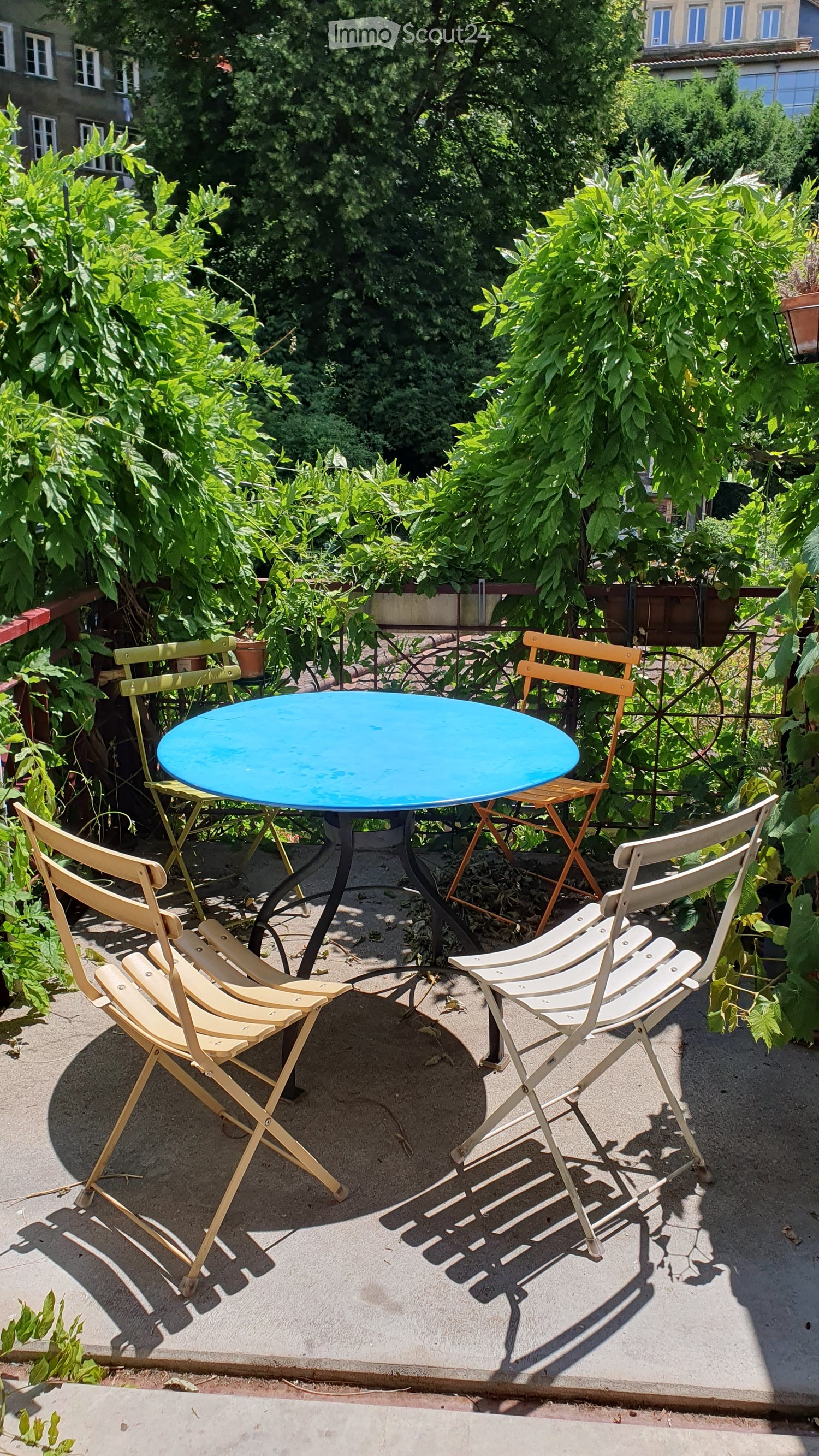 Balcony with a blue round table and several folding chairs surrounded by lush green plants and trees.