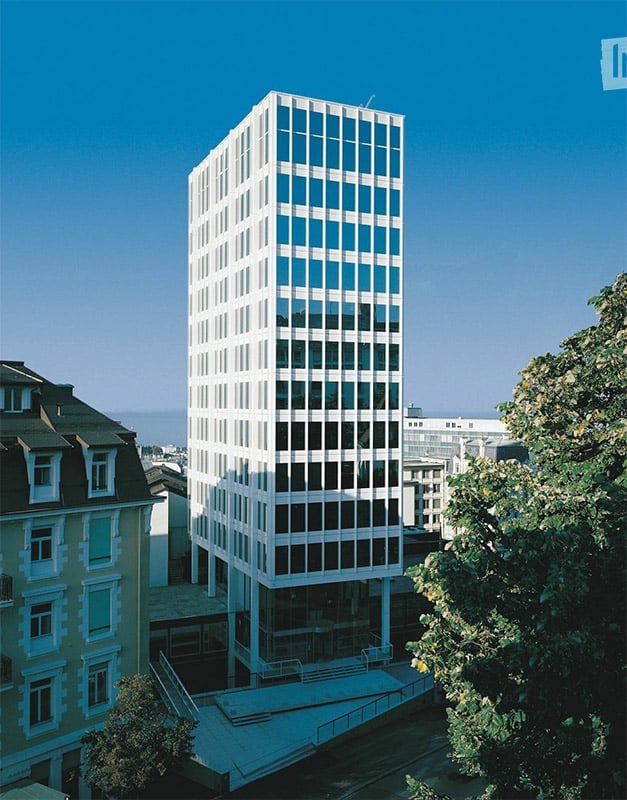 Modern high-rise building with a grid of windows, surrounded by smaller structures and greenery.