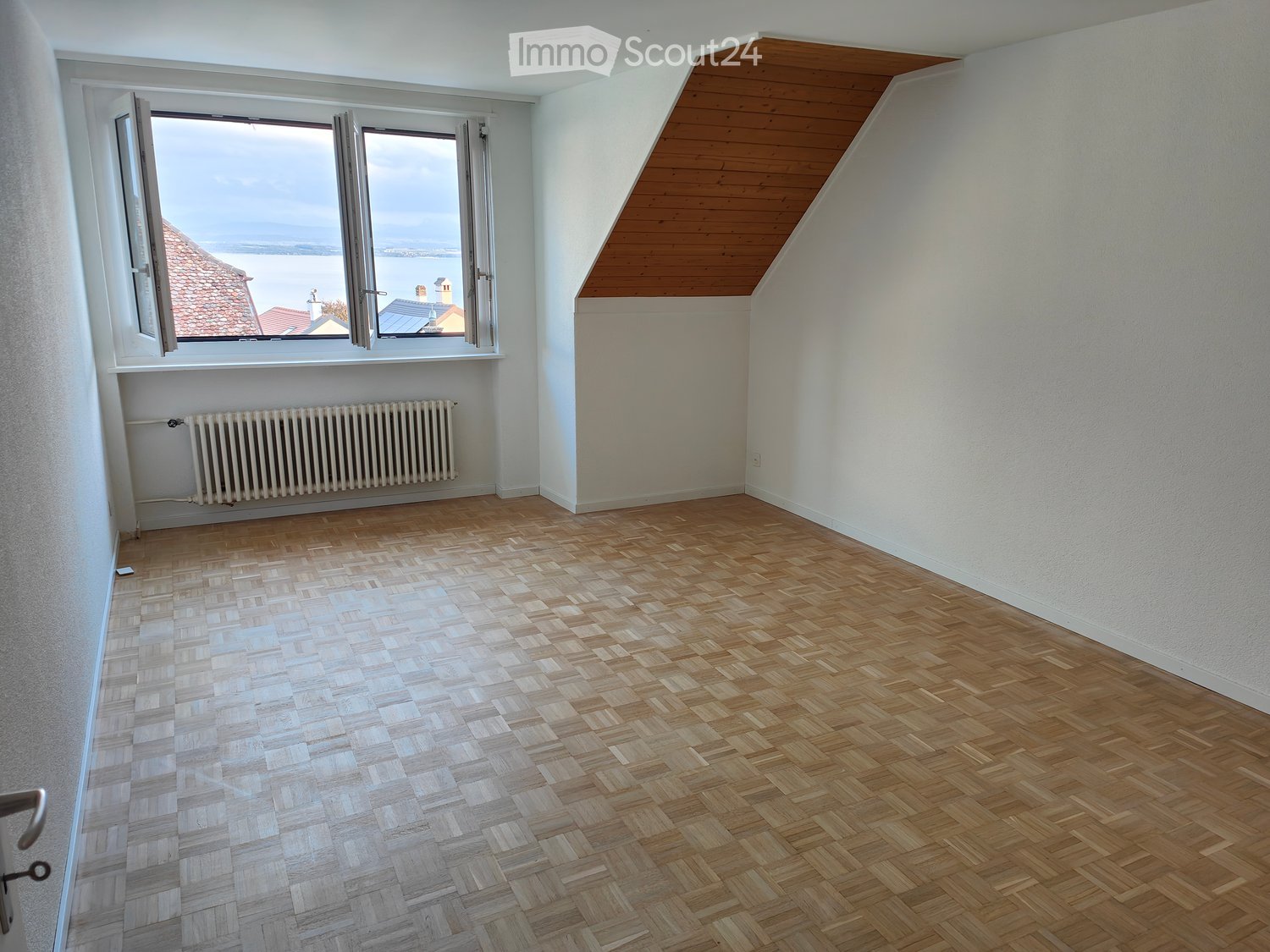 Empty room with wooden herringbone flooring, white walls, and a large window offering a view of the sea and distant rooftops. A radiator is mounted below the window.