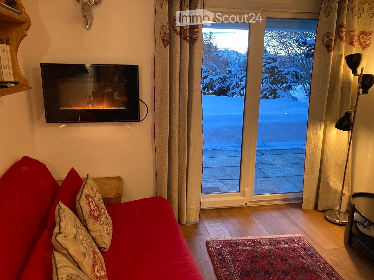 A cozy living room with a red sofa, decorative pillows, a wall-mounted fireplace, a floor lamp, a small table, and a shelf. Large windows offer a view of a snowy outdoor scene.