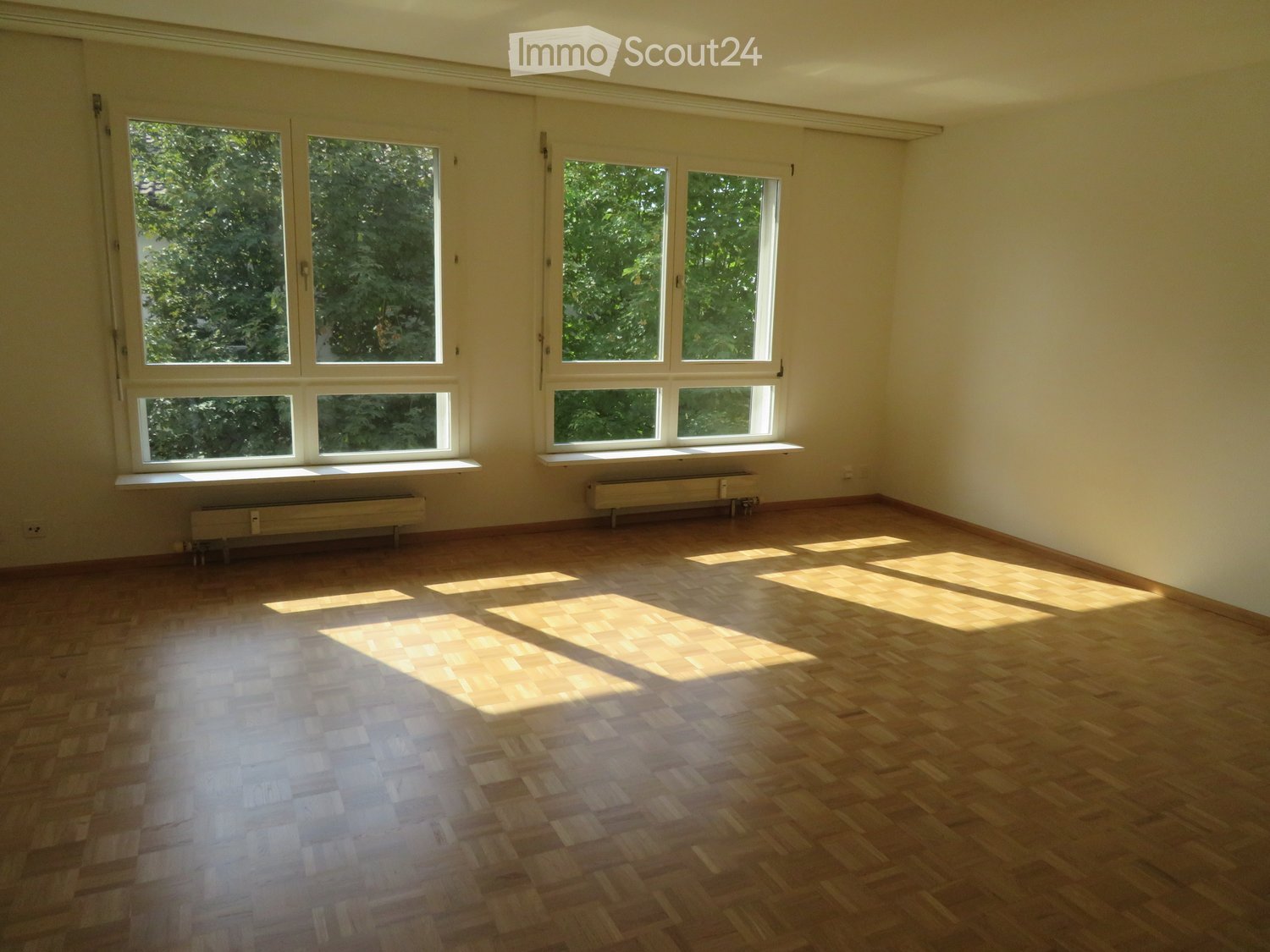 Empty room with wooden flooring and large windows allowing natural light to enter, with radiators beneath the windows.