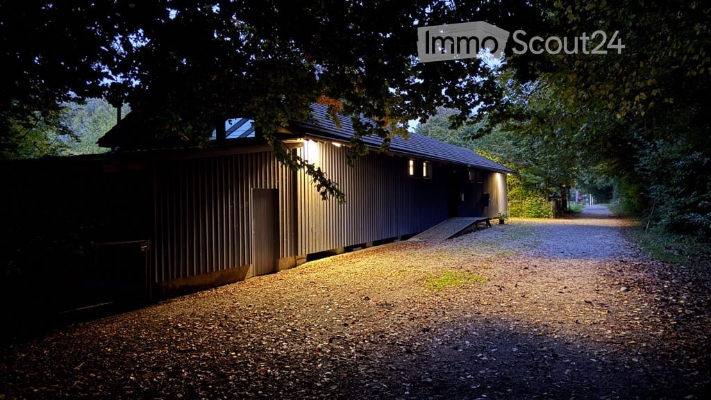 A modern, single-story building with a dark wooden facade and a gravel driveway, illuminated by warm lighting at dusk.