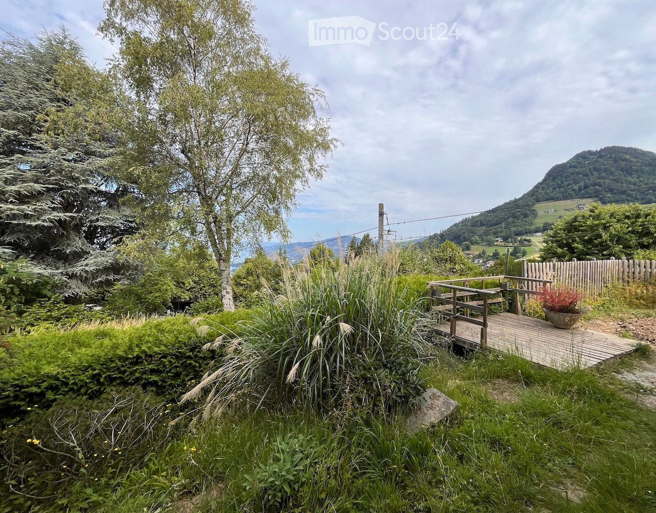 Wooden deck with a bench and potted plant, surrounded by lush greenery and trees, overlooking a scenic valley and hills.