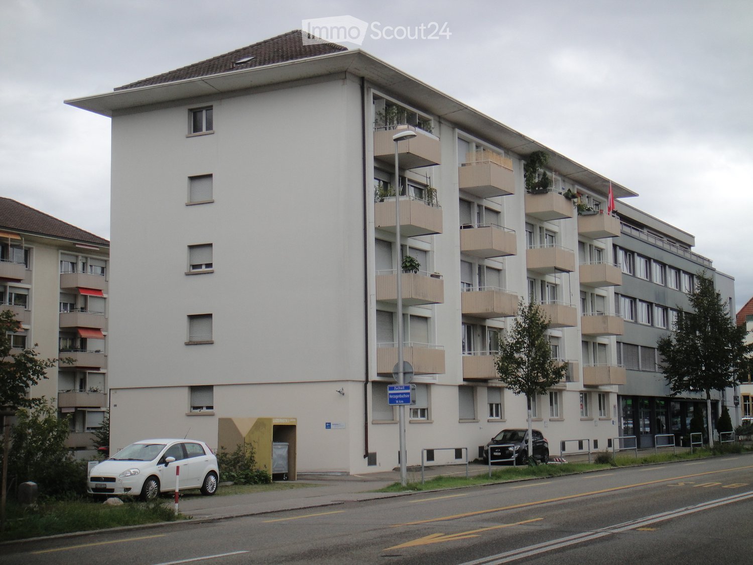 Four-story residential building with multiple balconies, white facade, and beige railings. Two cars are parked in front of the building.