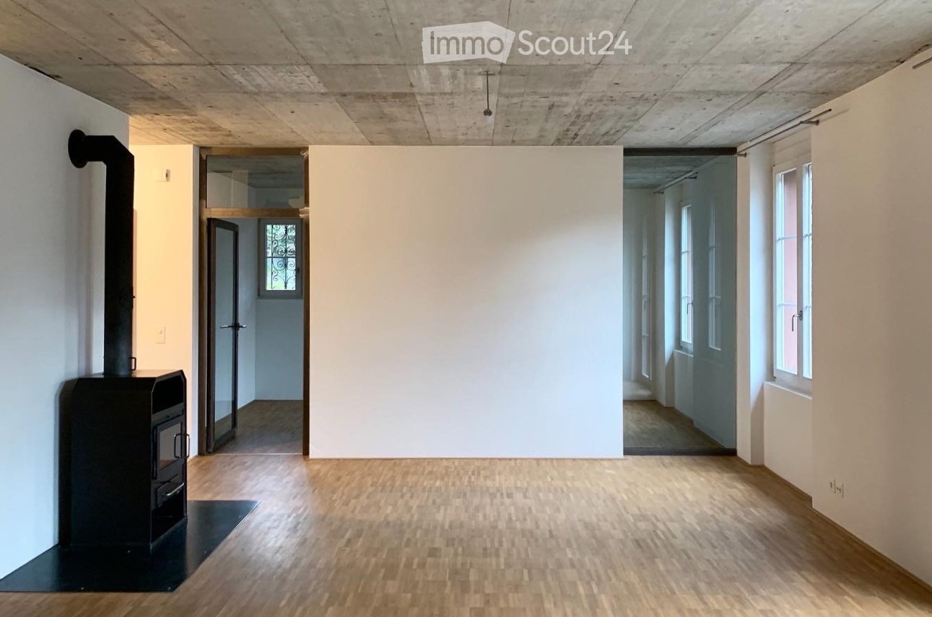 Empty room with wooden flooring, white walls, and a visible fireplace. The room has a doorway leading to another area and a window on the right side.