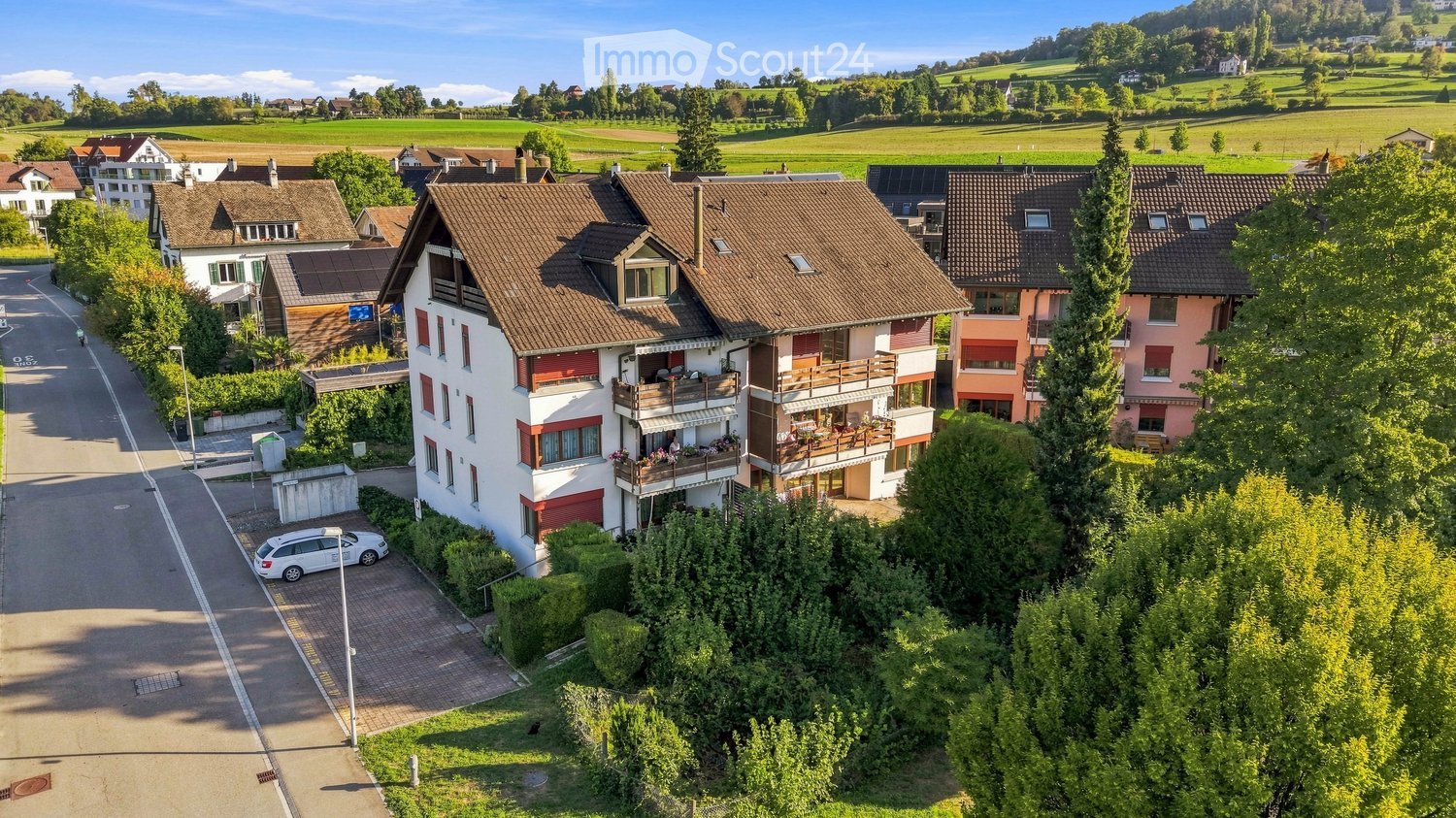 Aerial view of a residential area with multiple houses, balconies, and a parking area. The houses have red-tiled roofs and are surrounded by greenery and open fields.