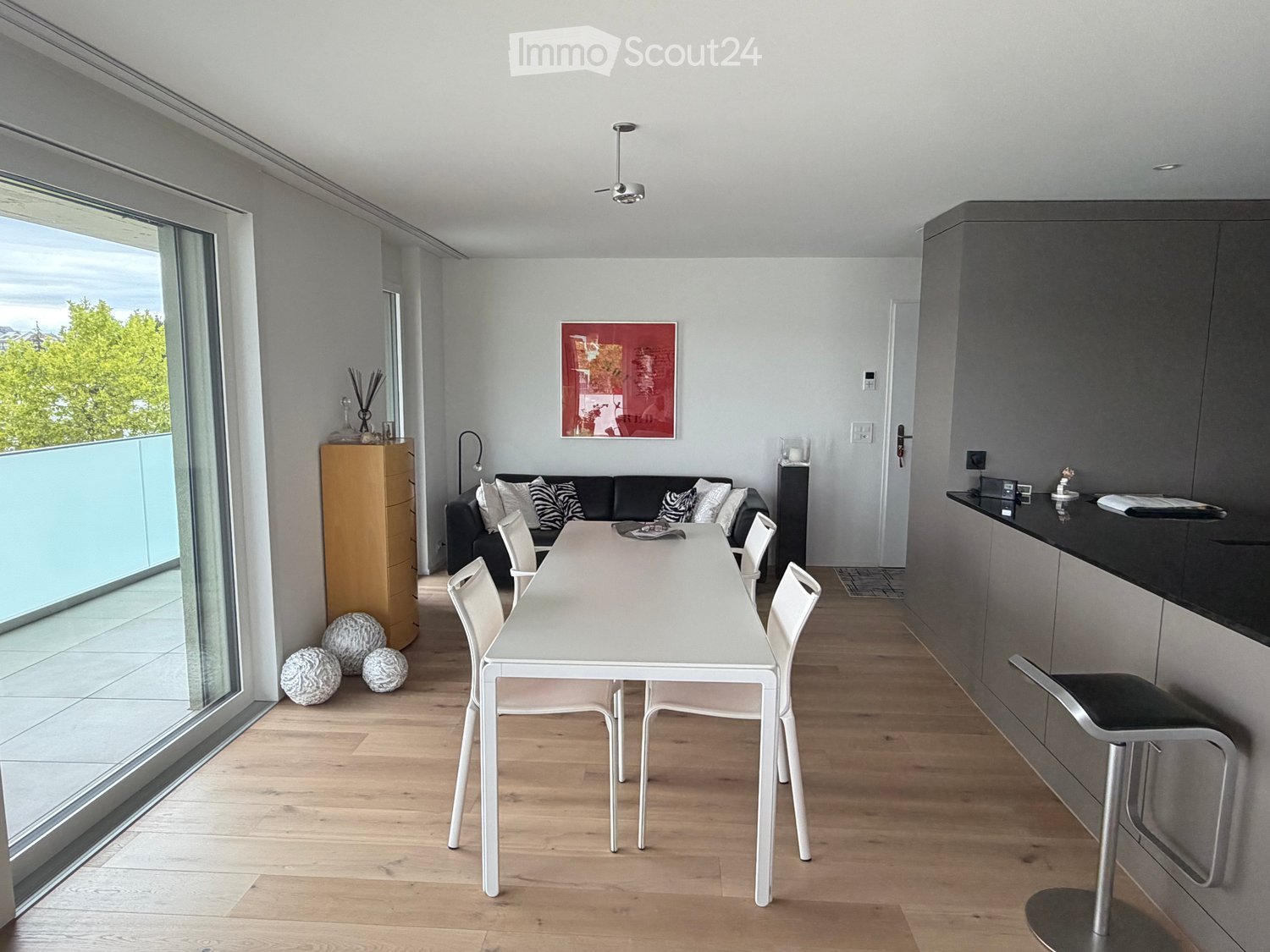 Modern living and dining area with a white dining table, chairs, a black sofa, a wooden cabinet, and a view of a balcony.
