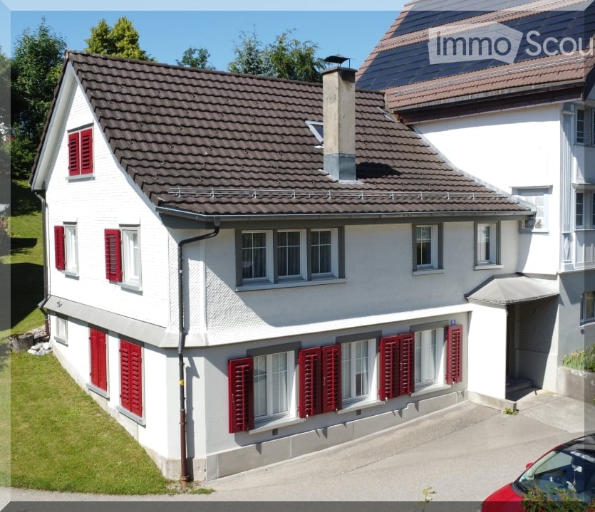 Two-story white house with red shutters, multiple windows, and a chimney. The house has a tiled roof and is surrounded by greenery.